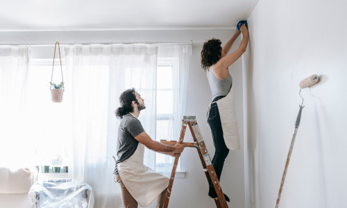 A couple prepping a wall with blue tape for paint.